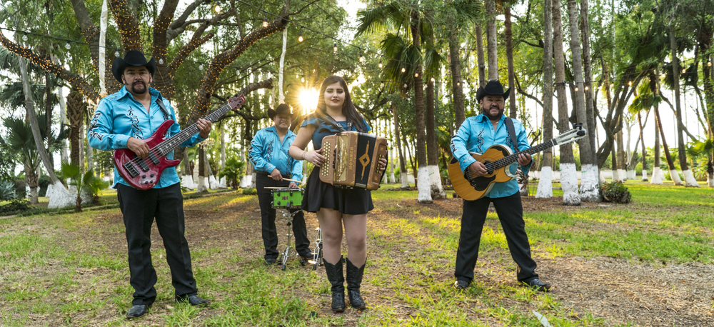 Regional Mexican band Grupo Control holding their instruments and playing in front of a setting sun in a park